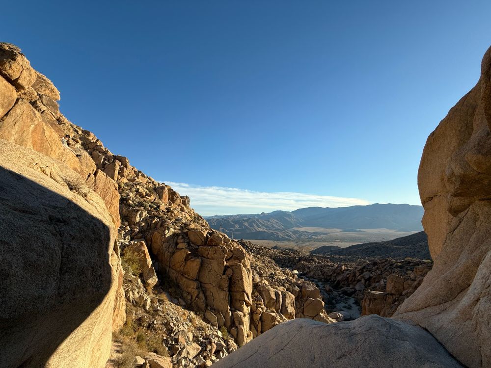A rocky hillside frames a beautiful desert valley view. Blue skies with a thin band of clouds fill the top half of the image. 