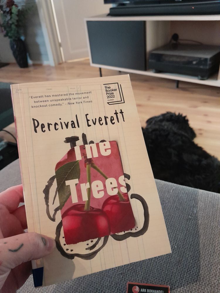 The Trees by Percival Everett, a couple of cherries on the cover, a dog lying on the floor behind the book