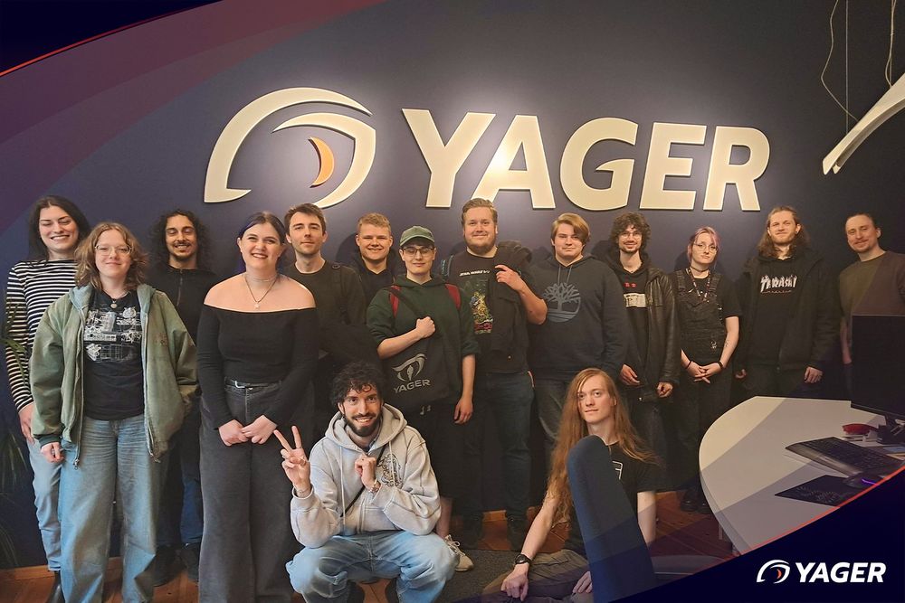 several people of different gender standing in an office in front of a YAGER logo
