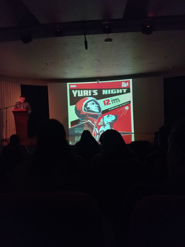 Una pantalla en un auditorio con un afiche rojo, blanco y negro que dice "Yuri's Night".
