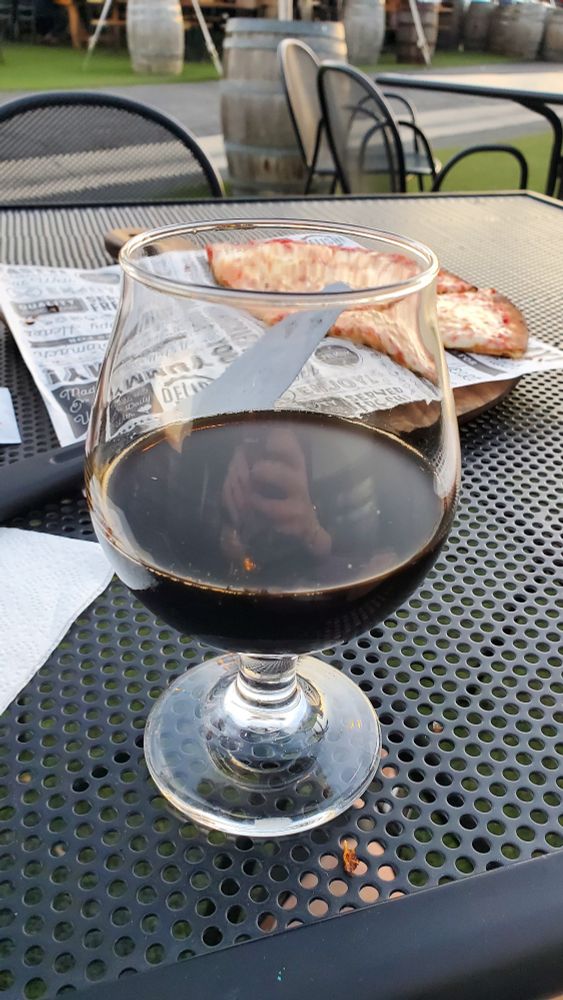 in an outside patio area of a local brewery, a glass of stout beer sits on a patio furniture table. in the background of the table is a half eaten cheese pizza. beyond the table, and out of focus, is fake lawn, beer barrel, etc making for a typical brewery type scenery.