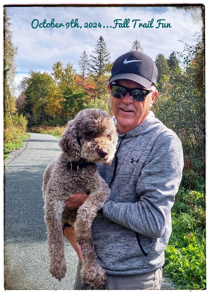 A man wearing sunglasses carrying a brown and gray dog. They are on a trail with vibrant fall colours and the background. 