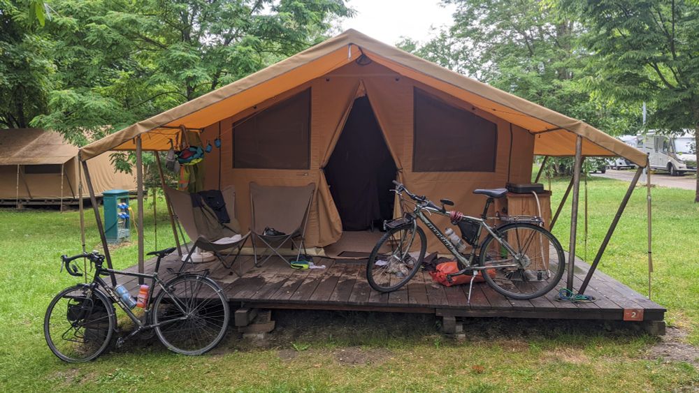 Two bikes in front of a large canvas tent. Another tent and a trailer can be seen in the background. 