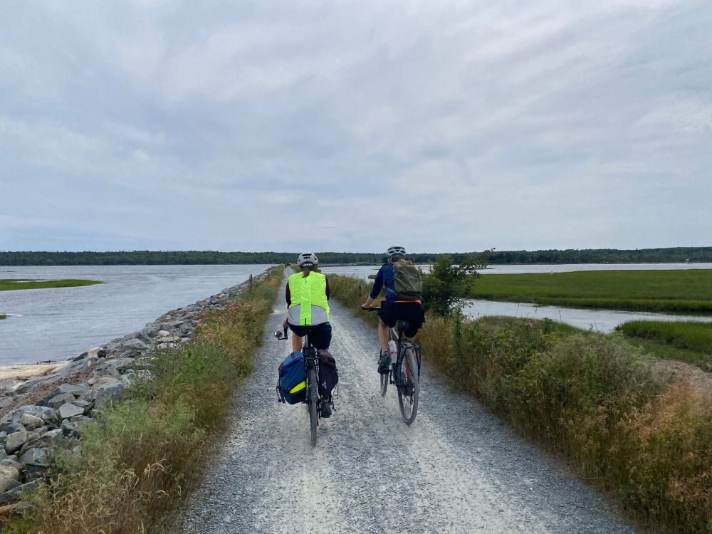 Two cyclists (V's daughters) riding on a gravel trail with grass and water on each side.
