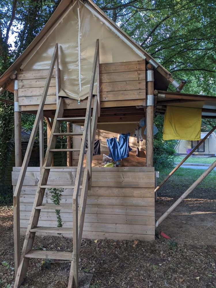 An elevated canvas tent with a steep set of stairs leading to the opening. There are clothes hanging on the lower level. 
