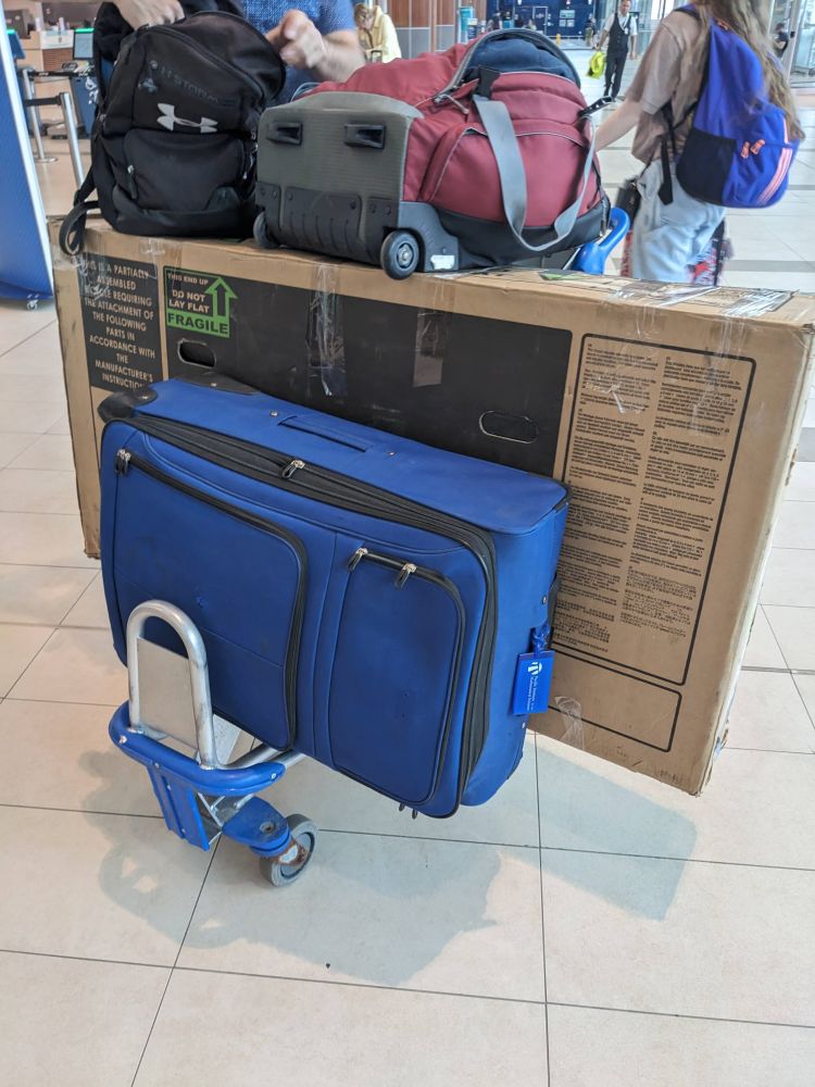 A large blue suitcase and a cardboard box with a bike in it on an airport luggage trolley. 