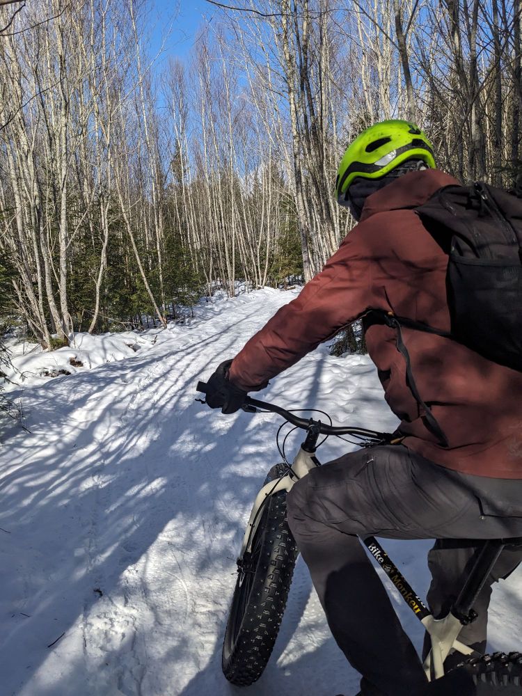 A cyclist on the side of a trail covered in snow on a sunny day. 