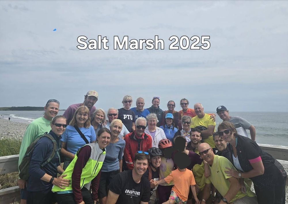 A group of about 25 people posing for a photo on a boardwalk with a beach in the background. The words "Salt Marsh 2025" are at the top of the photo in block letters.