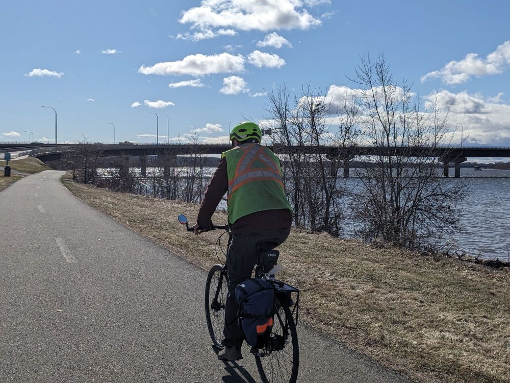 A cyclist in a yellow vest riding on a paved path next to a river. There is a bridge in the distance. 
