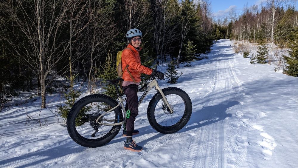 A cyclist wearing a bright orange coat standing over top of a fat bike on the side of a groomed trail. 