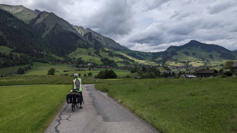 A cyclist in a yellow vest standing on a road with lush green mountains in the background. The sky is gray with thick clouds. 