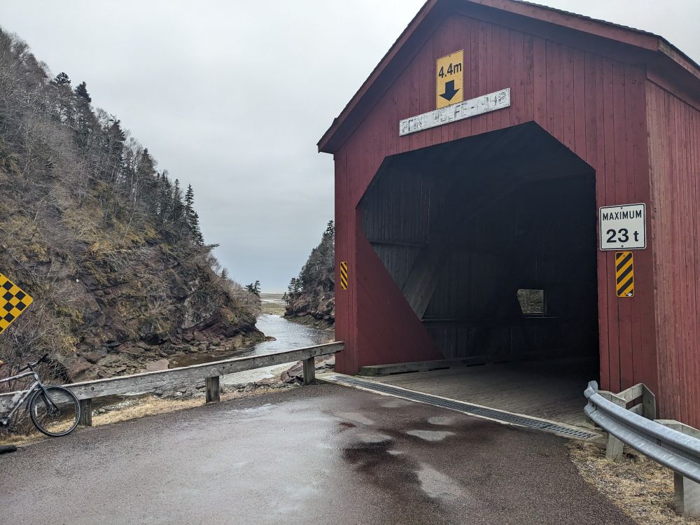 A red covered bridge with cliffs and a river to the left. 