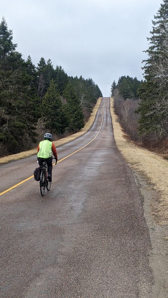 A cyclist in the middle of the road on an overcast day. A steep hill looms ahead of them. 
