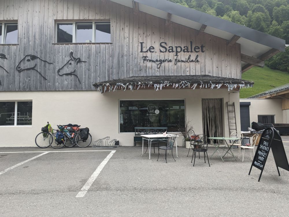 A fromagerie called Le Sapalet. There are images of goats and cows on the building and two bikes leaning on it. On the window there is text indicating "en self service" and the sign out front indicates that it is open 24/7.