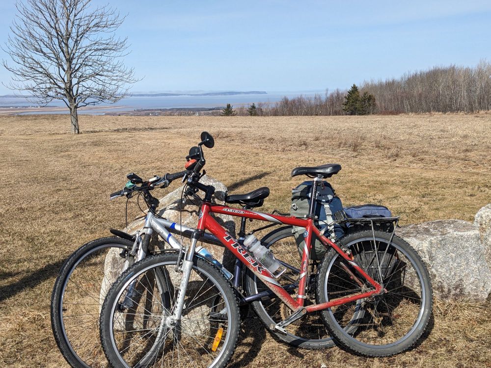 Two bikes leaning against a rock with a yellow field in the background. Blomidon can be seen in the distance. 
 