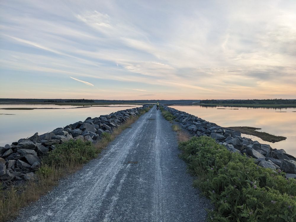 A gravel trail with boulders and water on each side. There is a cyclist in the distance. The sky has white and pinkish tones and the water is so still that it is acting like a mirror. 