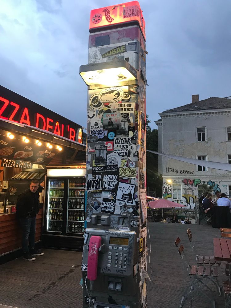 Mit Zetteln und Aufklebern zugeklebte Telefonsäule. Im Hintergrund eine Pizzeria mit Leuchtreklame.