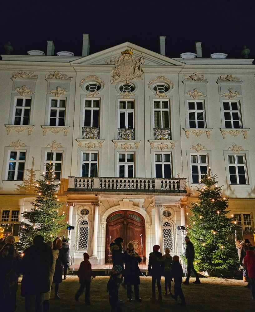 Weißes Schlossgebäude mit Holzportal, links und rechts Weihnachtsbäume.