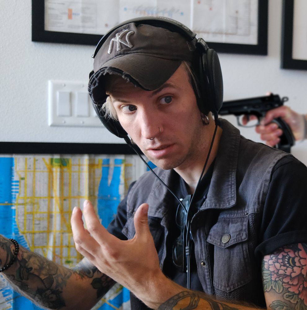 The author, a white man wearing a baseball cap, looking over his shoulder, distressed, as an offscreen person threatens him with a gun