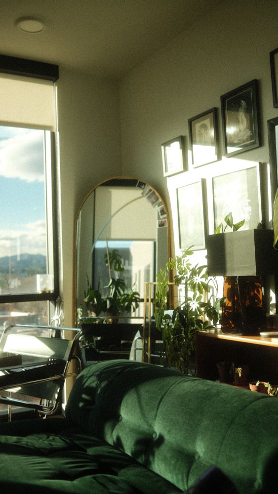 A corner author's living room, lit in a hazy, blooming sort of of afternoon light. In the furniture and features of the room are walnut, deep jeweled green, and amber acrylic tones, and the air is gold that you can touch.