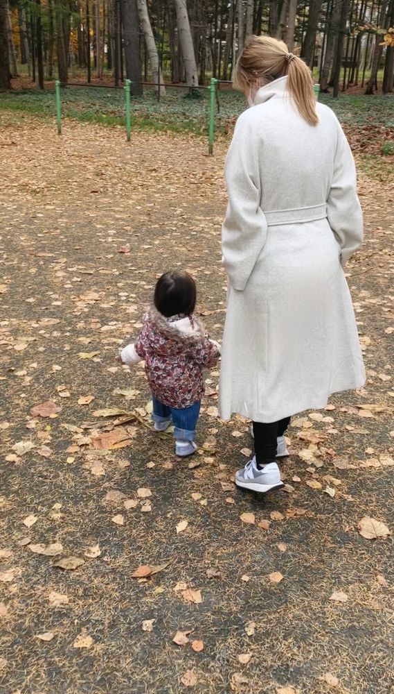 Ein kleines Kind mit dunklem Haar in einer dicken Jacke, das über einen Laub bedeckten Spielplatz läuft. Neben ihm eine Frau in einem weißen Mantel.