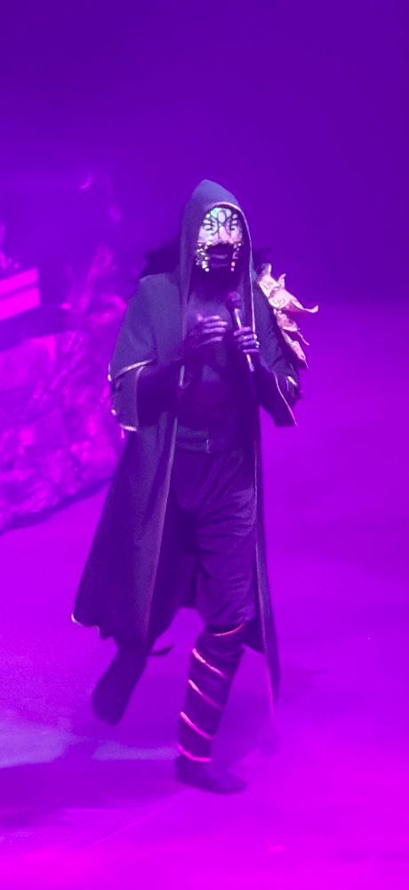 Vessel from Sleep Token, walking and singing. The lighting is a fuschia color and he’s walking towards the camera. 