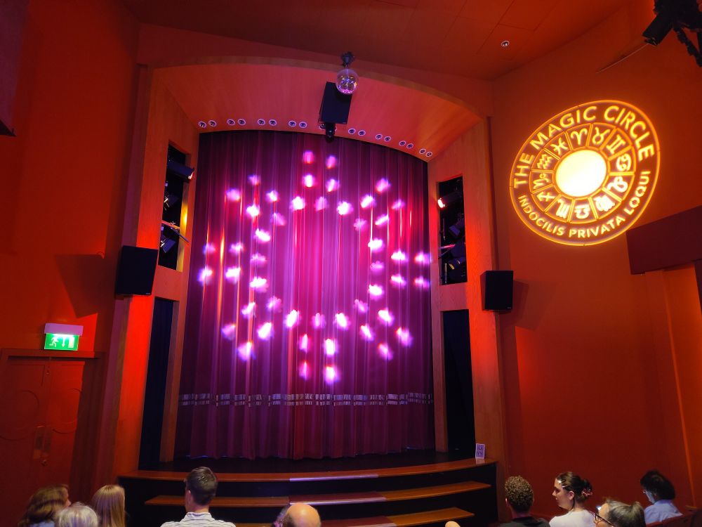 A red theatre, with curtain closed, and the magic circle emblem projected on the wall near the front.