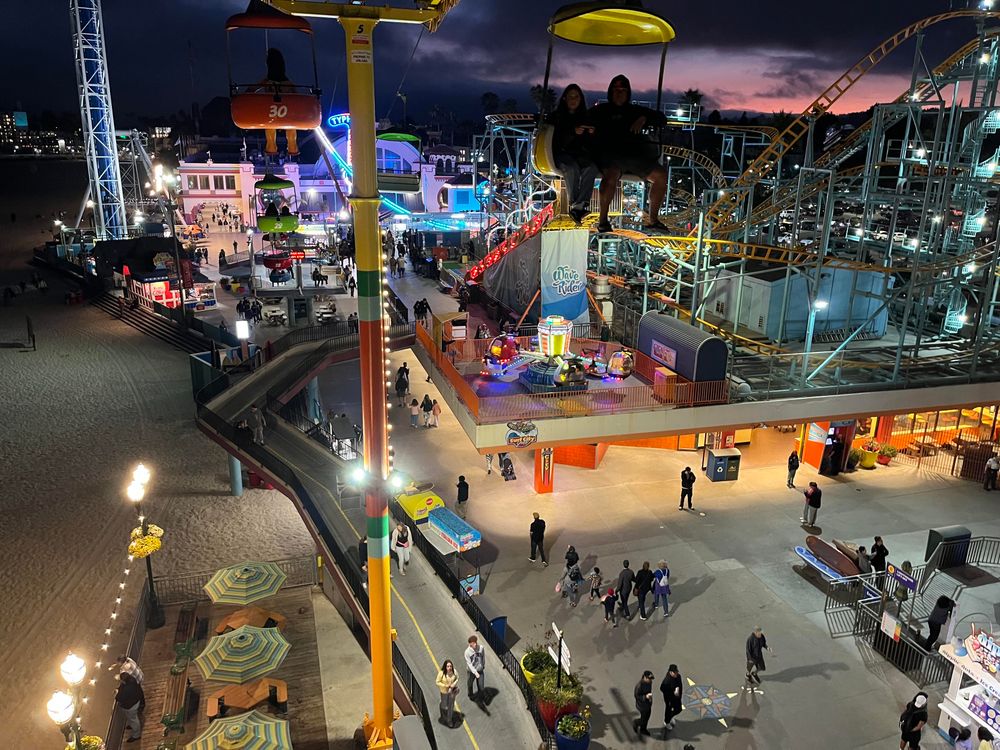 This is a beautiful evening photograph taken from a high vantage point at a seaside amusement park or pier. The image shows a classic carnival scene during twilight, with a dramatic purple and pink sunset sky in the background.

In the foreground, there’s a large yellow Ferris wheel support structure, and you can see gondolas with passengers enjoying rides against the colorful sky. The scene below reveals a bustling boardwalk filled with people walking around, illuminated by colorful carnival lights and attractions.

The setting is coastal, with a beach visible, and palm trees silhouetted in the distance. The warm carnival lighting creates a nostalgic, magical atmosphere as day transitions to night.
Various carnival rides and attractions are visible throughout the scene, including games and food stands. The photograph captures that special golden hour moment when amusement parks come alive with lights and activity, creating a festive and enchanting atmosphere.​​​​​​​​​​​​​​​​