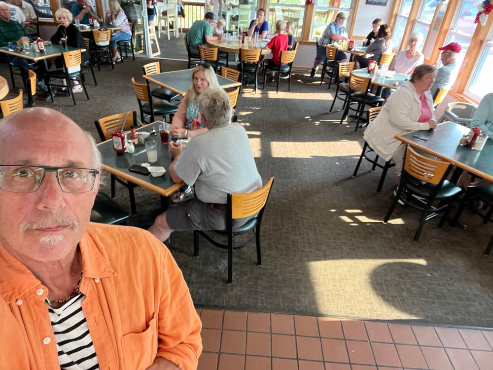 This photo shows the interior of a casual restaurant with a warm, welcoming atmosphere. In the foreground, a man [it’s me!] wearing glasses, an orange button-up shirt, and a black and white striped shirt underneath is taking a selfie. He has a gray beard and mustache.

The dining area features numerous wooden tables with matching wooden chairs that have horizontal slats in their backs. The floor appears to be tiled, and there’s carpeting in the seating area. Large windows along one side let in natural light, creating bright patches of sunlight on the floor.

The restaurant appears busy with diners of various ages seated throughout the space. You can see people engaged in conversation at their tables, with drinks and table settings visible. The overall ambiance suggests this is a popular local dining spot, possibly during lunch or brunch hours given the bright natural lighting streaming through the windows.

The décor and furniture style gives the restaurant a family-friendly, neighborhood establishment feel rather than fine dining - the kind of place where people come for comfort food and casual meals.​​​​​​​​​​​​​​​​