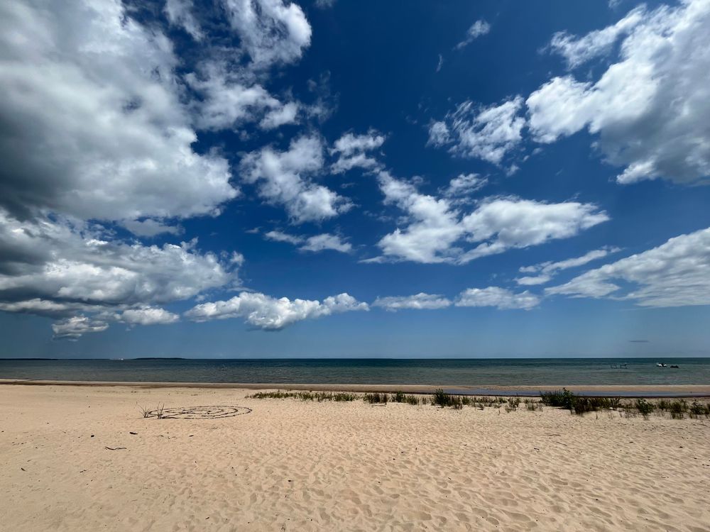 This photo shows a beautiful beach scene with pristine sandy shores stretching toward calm blue-green waters. The sky dominates much of the frame, filled with dramatic white and gray clouds against a deep blue backdrop. The beach appears relatively undeveloped, with natural dune grass growing behind the shoreline and what looks like a simple circular pattern or marking drawn in the sand in the foreground. Small boats or objects can be seen on the distant horizon. The lighting suggests it’s a partly cloudy day with good visibility, creating an appealing contrast between the white sand, blue water, and dynamic cloudscape above.​​​​​​​​​​​​​​​​