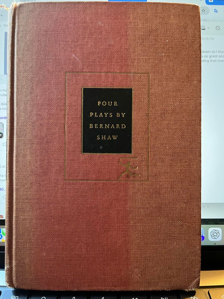This photo shows the cover of a vintage hardcover book titled “Four Plays by Bernard Shaw.” The cover is a textured reddish-brown cloth with a black rectangular label centered near the top. The title and author’s name are stamped in gold lettering on the label:

FOUR  
PLAYS BY  
BERNARD  
SHAW

A gold-inked rectangular border surrounds the black label, and at the bottom right of the border is a small, stylized design that resembles a publisher’s colophon. The cover shows some wear, indicating that the book is likely an older edition.