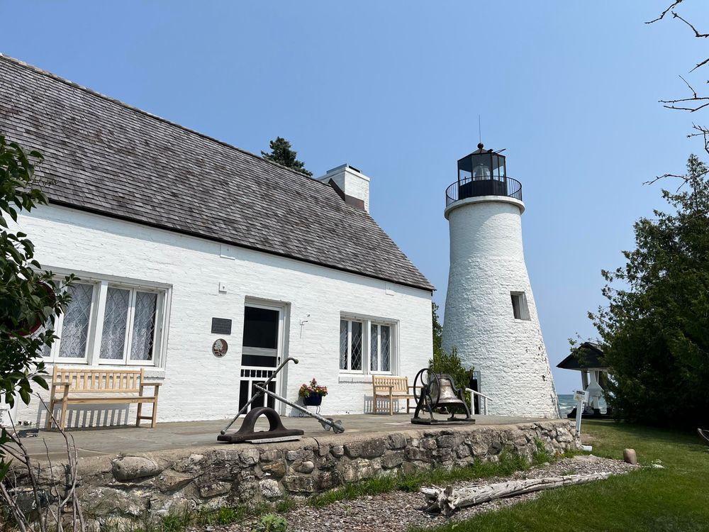 These photo shows a shorter, more conical white lighthouse with a black lantern room at the top. This lighthouse is built near a rocky shoreline, as evidenced by the stone wall or foundation visible in the foreground. The associated building is a white house with a distinctive slate or shingle roof, and there are wooden benches positioned around the property. An old anchor is displayed on the stone wall, adding to the maritime heritage theme. The setting appears more coastal and rugged compared to the first lighthouse.