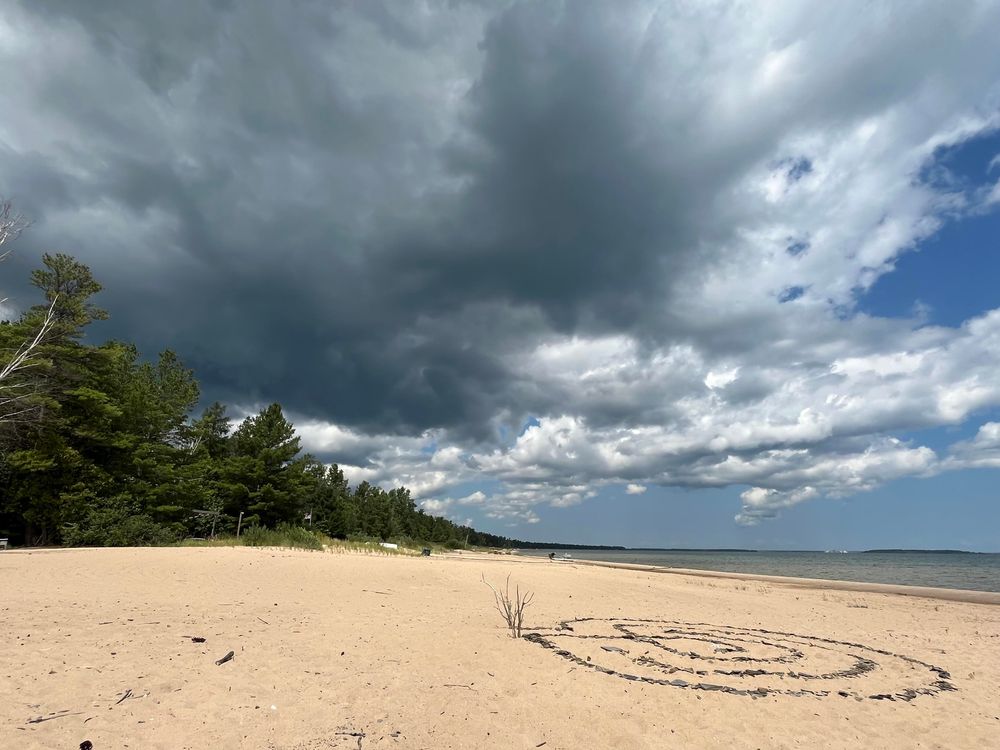This photo captures the same beach location but with dramatically different weather conditions. The sky is now dominated by dark, ominous storm clouds that create a moody, overcast atmosphere. The lighting is much more subdued and gray compared to the bright, sunny conditions in the previous images. You can see the same spiral pattern in the sand, though it appears less distinct in this lighting. The beach stretches into the distance where there’s a tree line visible on the left side of the frame. The contrast between the dark, threatening sky and the pale sand creates a striking atmospheric effect, suggesting an approaching storm or unsettled weather conditions. The overall mood of this image is much more dramatic and foreboding than the earlier sunny beach scenes.​​​​​​​​​​​​​​​​