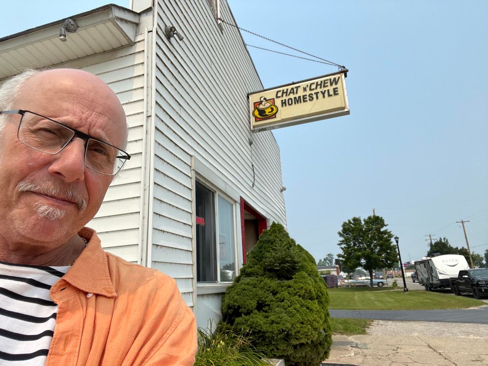 This photo shows a man [it's me!!] taking a selfie in front of a small, local restaurant. He's wearing glasses, has a gray goatee, and is dressed in an orange/coral colored shirt or jacket over what appears to be a striped shirt underneath.

Behind him is a white building with horizontal siding that houses "Chat 'N Chew Homestyle" restaurant, as indicated by the hanging sign with a coffee cup logo. The building is a converted residential structure or small commercial building typical of family-owned establishments in smaller towns or rural areas.

The setting is a highway with power lines visible, some RVs or trailers parked in the background, and well-maintained landscaping including a nicely shaped evergreen shrub in front of the restaurant. The sky is clear and blue, suggesting it's a pleasant day for dining out or exploring local eateries.

This appears to be the kind of homestyle restaurant that travelers and locals might seek out for comfort food and a friendly atmosphere. 