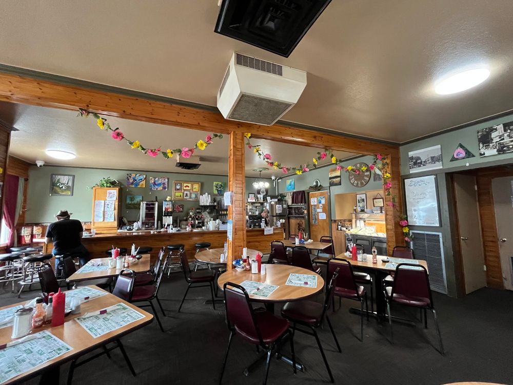 This photo shows the interior of a cozy, small-town restaurant. The atmosphere is warm and homey, with wood-paneled pillars and trim, dark carpeted floors, and simple furnishings.

Several tables with maroon chairs are arranged throughout the dining area. Each table has laminated menus, ketchup and mustard bottles, salt and pepper shakers, and sugar dispensers. The restaurant is decorated with colorful artificial flower garlands strung across the ceiling beams, adding a festive and cheerful touch.

In the background, there’s a counter area with barstools and a person sitting at the counter. Behind the counter, you can see coffee machines, drink dispensers, and various items for food service. The walls are decorated with framed pictures, local artwork, and vintage photographs. A large wall clock and a dry-erase board listing “Specials” are also visible, along with a dessert display case.

The overall vibe suggests a friendly, down-to-earth diner popular with locals.