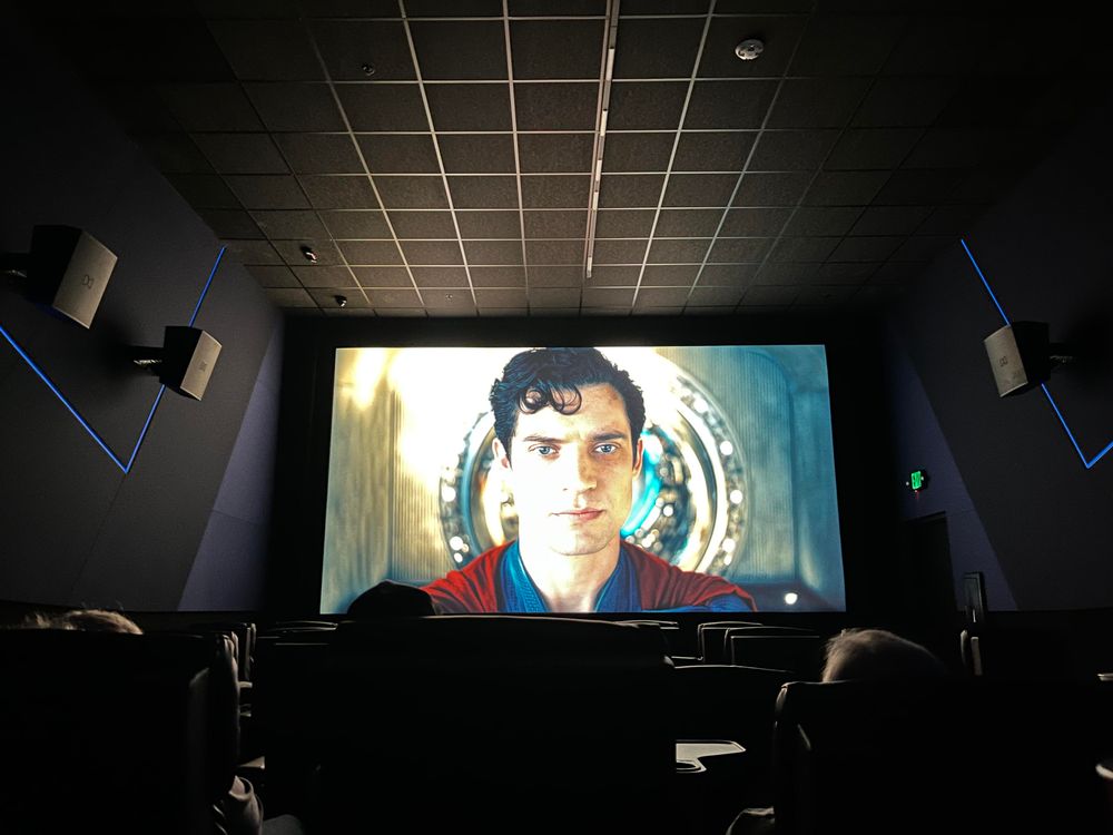 This photo shows the interior of a modern movie theater. The image is taken from the perspective of a seated viewer, looking toward the large screen at the front of the auditorium. On the screen, a close-up of a young man in a red garment is shown; he has dark, curly hair and is looking directly at the camera. [It’s Superman, you stupid robot!] The background of the scene on the screen features a circular, metallic, futuristic design.

The theater itself has reclining seats, cup holders, and dark walls with blue LED accent lighting running diagonally across the walls. Surround-sound speakers are visible on the side walls. A green exit sign is illuminated near a door on the right side of the room. A few heads of other moviegoers are visible in the bottom portion of the image, silhouetted against the light from the screen.