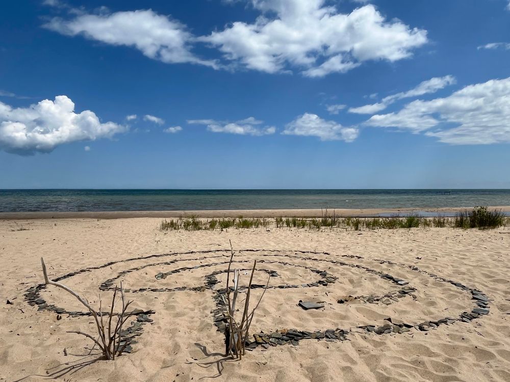This photo shows the same beach location from a closer perspective, focusing on an intricate spiral pattern in the sand made from dark stones arranged in concentric circular paths forming a walking meditation labyrinth. A few dried plant stems or branches stick up from within the pattern. The beach setting remains consistent with the first image - sandy shores, dune grass in the background, and the same blue-green ocean waters under a partly cloudy sky. This appears to be someone’s artistic creation or spiritual practice space on the beach, with the labyrinth carefully constructed as a temporary sand art installation.​​​​​​​​​​​​​​​​