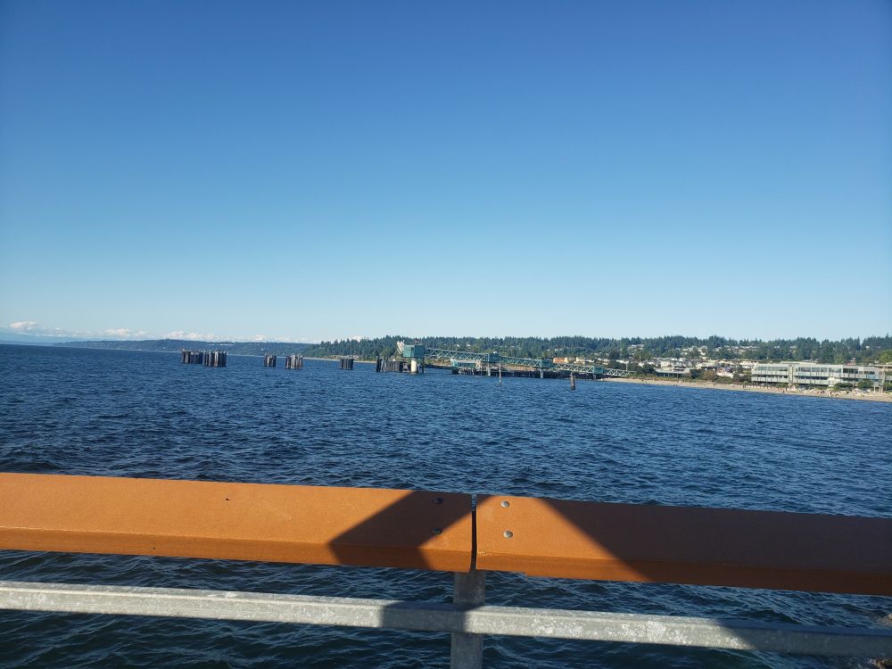 View from a pier over the Puget Sound. The sky is cloudless, the water a dark blue. The Edmonds coastline is in the background. A wooden handrail runs along the foreground.