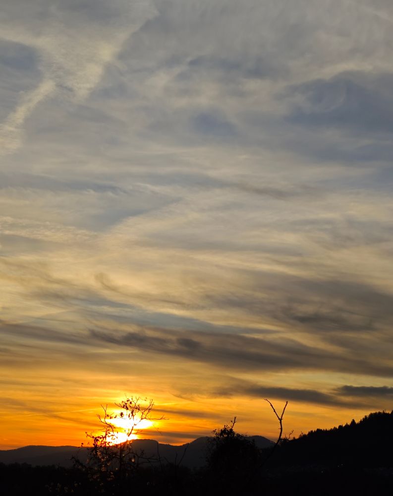 Das Bild zeigt einen Sonnenuntergang hinter einer Hügelkette. Der Himmel ist von unterschiedlich dichten Wolken bedeckt, die in Schichten und Strukturen verlaufen. Im Vordergrund sind Silhouetten von Bäumen und Ästen zu erkennen. Die Sonne steht tief am Horizont und beleuchtet die Wolken in hellen und dunklen Farbtönen.