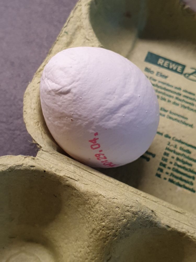 Foto: ein weißes Hühnerei pinkem Zahlenaufdruck in einem grünen Eierkarton. Die Eierschale sieht "runzelig" aus.