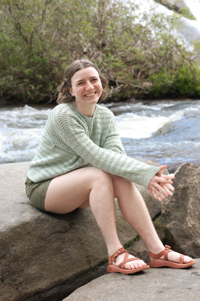 Andrea sits on a large rock by a creek wearing a light green handknit sweater. The sweater has stripes of eyelet lace and a relaxed fit. The sweater is styled with forest green shorts and brown Chacos.