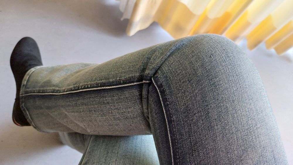 Das Bild zeigt die Beine einer Person, die Jeans und schwarze Schuhe trägt. Die Beine sind übereinander geschlagen. Im Hintergrund ist ein gelblich-weißer Vorhang zu sehen.
