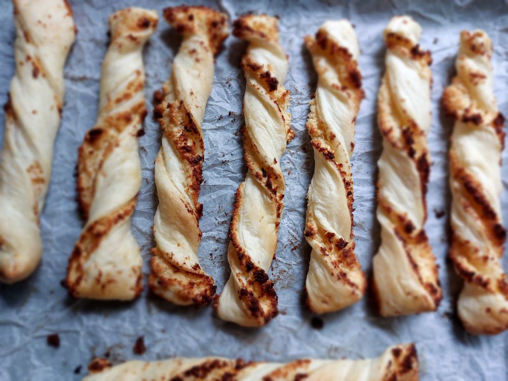 Das Bild zeigt eine Nahaufnahme von Kokos-Twists, die auf einem Backpapier liegen. Die Kokos-Twists sind goldbraun gebacken und haben eine spiralförmige Form. Das Backpapier ist leicht zerknittert und hat einige Krümel. Das Bild hat einen leicht unscharfen Hintergrund.