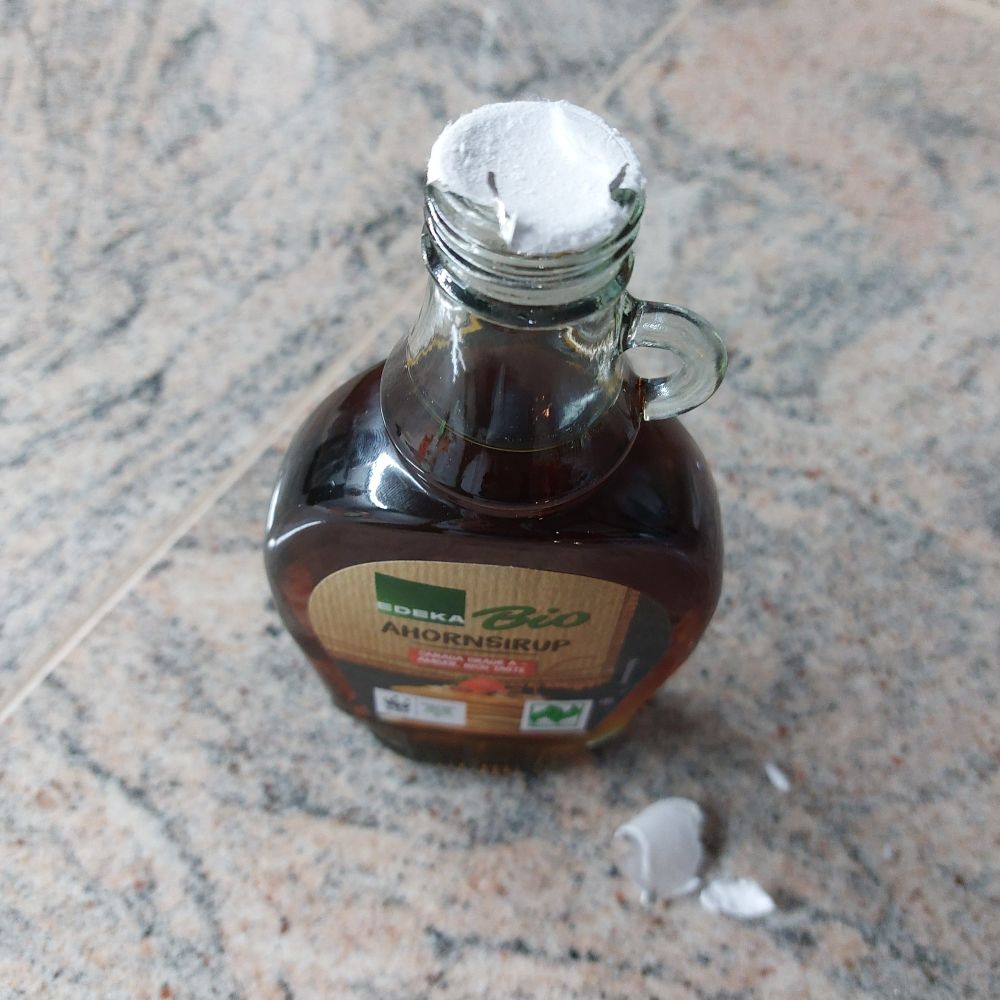 Das Bild zeigt eine Flasche Ahornsirup mit zum Teil abgefriemeltem Siegel. Teile davon liegen rechts vor der Flasche auf der Arbeitsplatte.