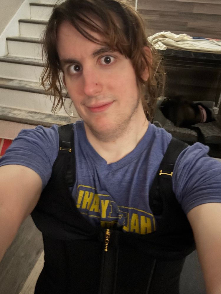 girl wearing black shapewear and smiling. her brown hair is pulled up into a bun. she’s wearing a blue tshirt and sitting on a gray couch. stairs and a dog are in the background. 