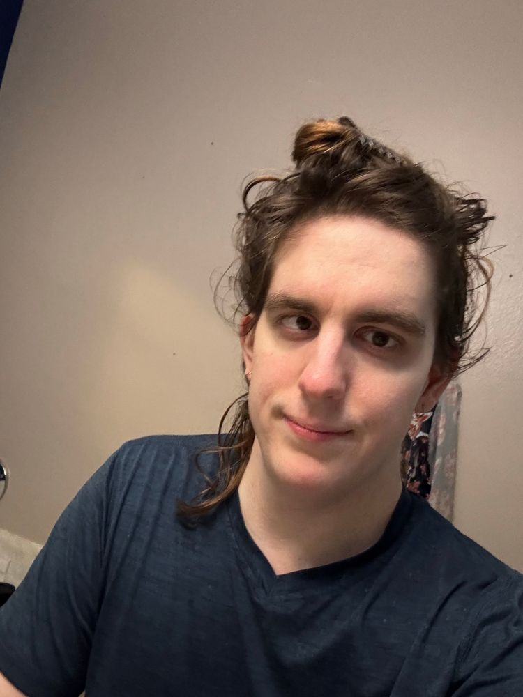 woman in a blue sporty t shirt. her face is smooth and freely shaved. brunette hair is up in a messy bun. she has small earrings and brown eyes. she’s smiling 