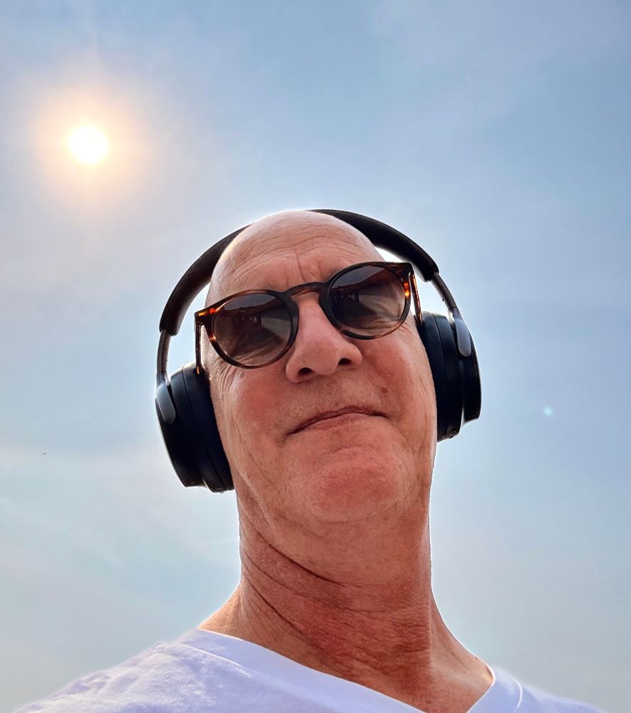 Selfie von Kopf und Hals mit Kopfhörern und Sonnenbrille im Gegenlicht und der Sonne als kleiner Ball am blauen Himmel.