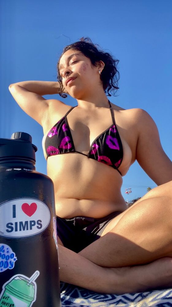 Skye is sitting on the beach legs crossed  stretching. There is a water bottle in the corner of the frame with a big sticker saying “ I ❤️ simps” 
