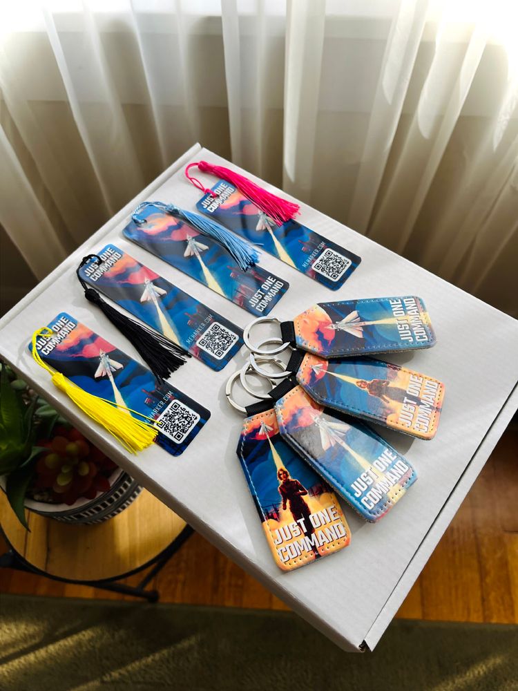 Several bookmarks and keychains are laid out on top of a box. They feature space-themed designs with rocketships, and have the book title "JUST ONE COMMAND" on them.