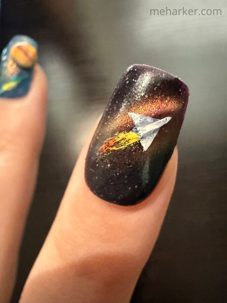 A detail shot of a space-themed painted nail. It features a rocket ship over a sparkly, outer-space background.